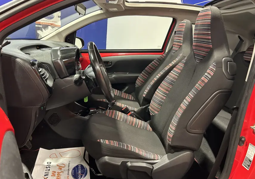 Vue intérieure côté conducteur d'une Citroën C1 rouge 2019, sièges tissu rayé noir et rouge, tableau de bord compact.