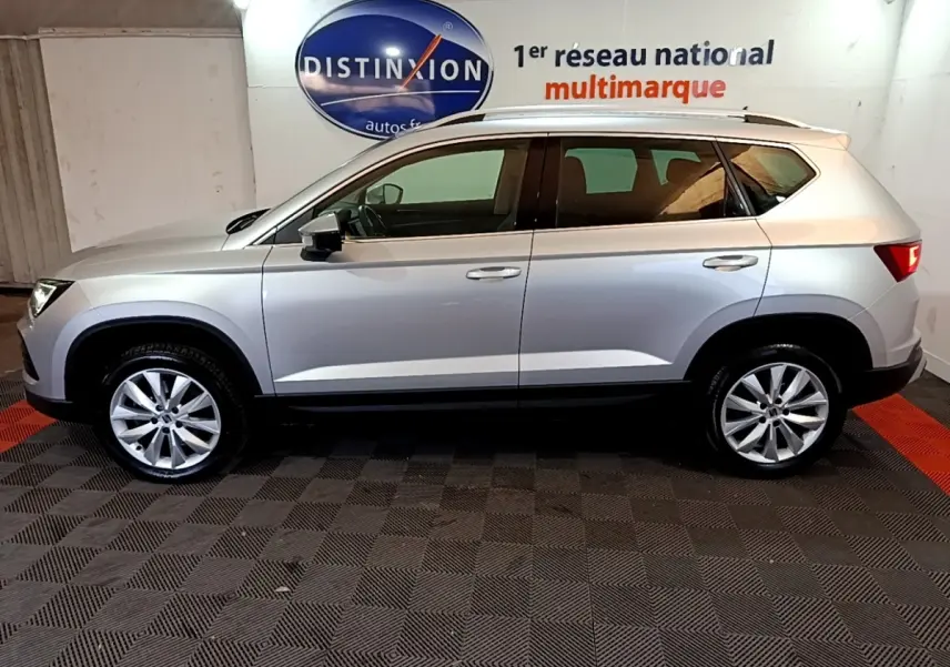 Vue de profil côté gauche d’un SEAT Ateca gris clair, avec jantes alliage et barres de toit chromées, en intérieur showroom.
