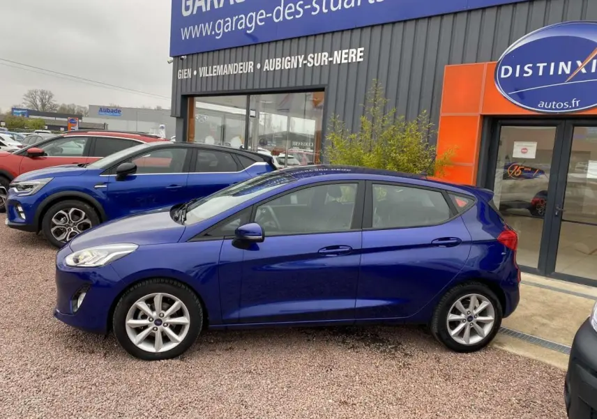 Vue de profil côté gauche d'une Ford Fiesta 1.1i Trend bleu foncé garée devant un garage, sur sol gravillonné.
