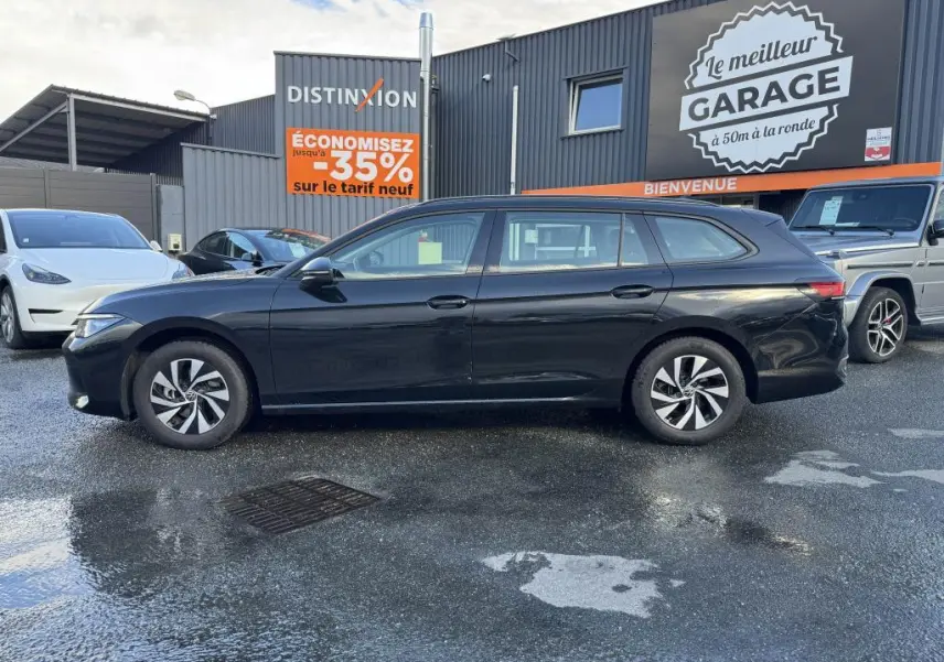 Volkswagen Passat SW noire vue de profil côté gauche, garée devant un garage avec panneaux publicitaires.