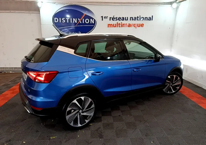 Vue 3/4 arrière droite d’un SEAT Arona bleu 2022 avec toit noir et jantes alliage distinctives en intérieur showroom.