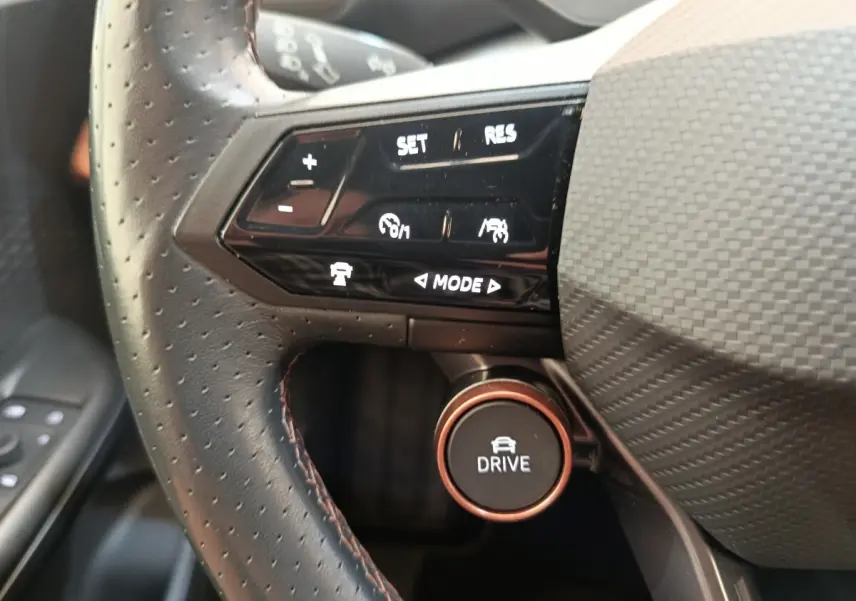 Gros plan sur les commandes du volant cuir noir perforé du Cupra Born 230ch, bouton Drive et réglages du régulateur.