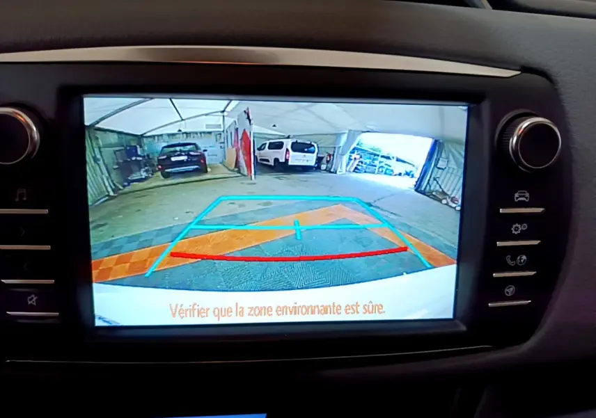 Écran tactile intérieur montrant la caméra de recul avec vue arrière d’un parking sous tente, Toyota Yaris blanche.