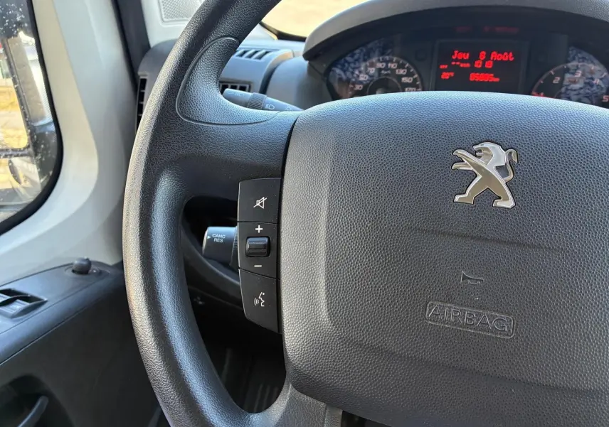 Gros plan sur le volant noir du Peugeot Boxer 2019 avec commandes intégrées et tableau de bord digital rouge en arrière-plan.