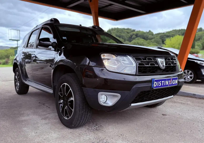 Dacia Duster noir métal en 3/4 avant droit, avec jantes bi-ton et calandre distinctive sous abri.