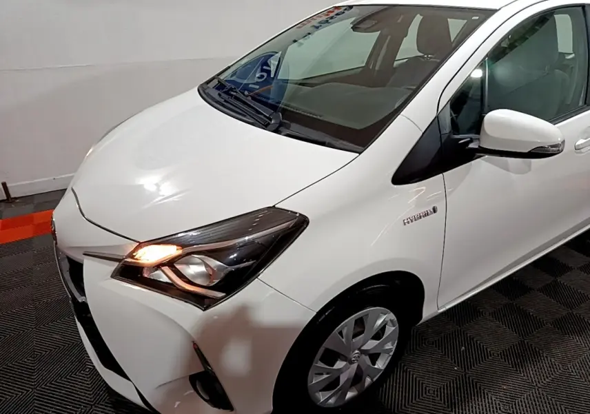 Vue 3/4 avant droit d'une Toyota Yaris blanche hybride avec feu avant allumé et logo Hybrid visible sur l'aile.