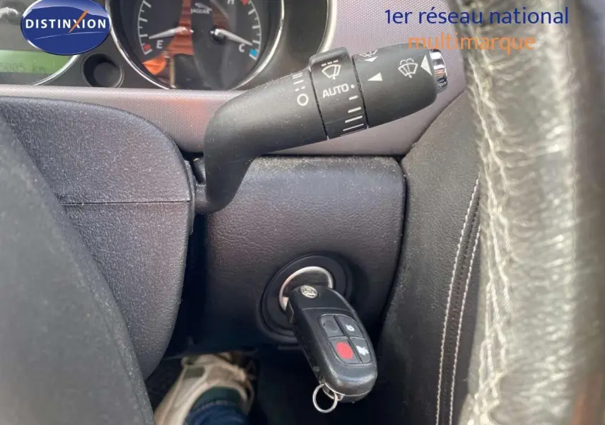Gros plan intérieur sur le contacteur avec clé Jaguar insérée, volant cuir noir visible en partie à droite.