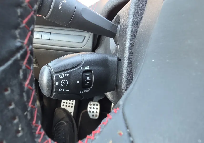 Gros plan sur la commande de régulateur de vitesse à gauche du volant cuir noir avec surpiqûres rouges dans une Peugeot 208 GTI noire.