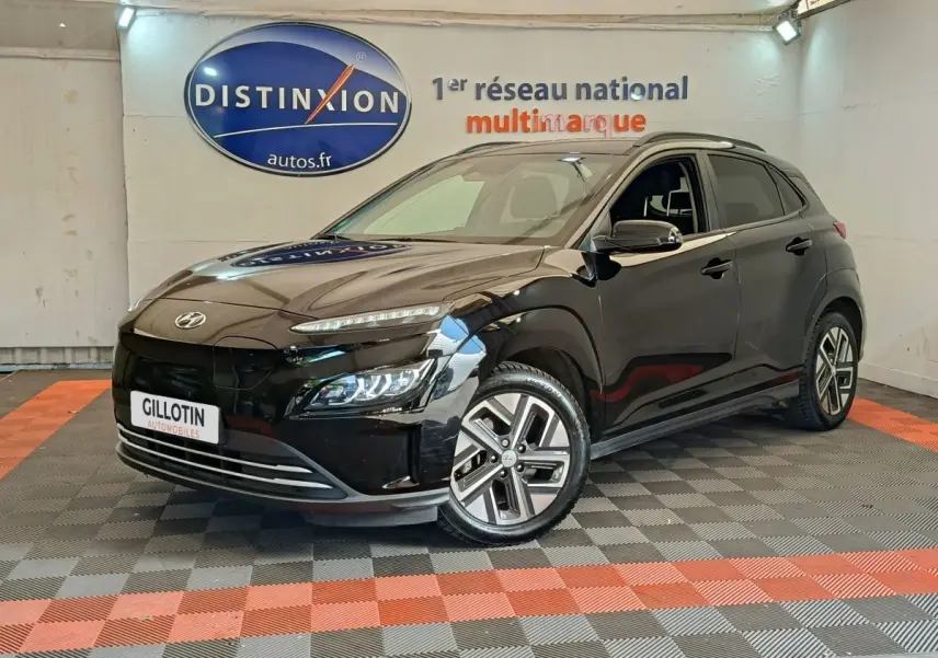 Hyundai Kona électrique noir en 3/4 avant droit, avec jantes spécifiques et feux LED allumés en intérieur showroom.