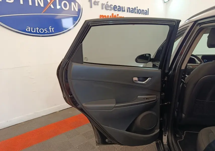 Vue intérieure de la porte arrière gauche ouverte d'une Hyundai Kona noire, avec siège arrière et tableau de bord partiellement visibles.