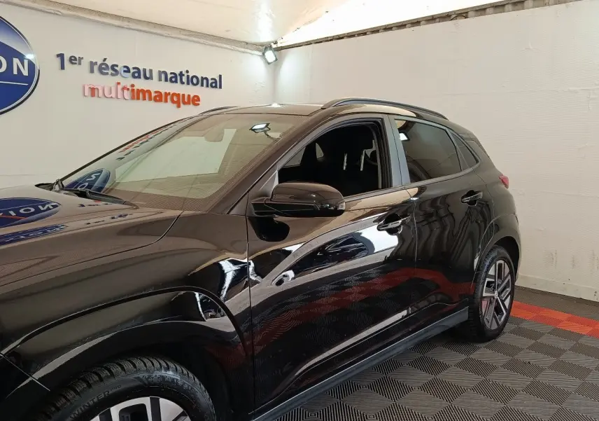 Vue côté gauche du Hyundai Kona électrique noir 2023 dans un showroom avec sol à motifs gris et rouge.