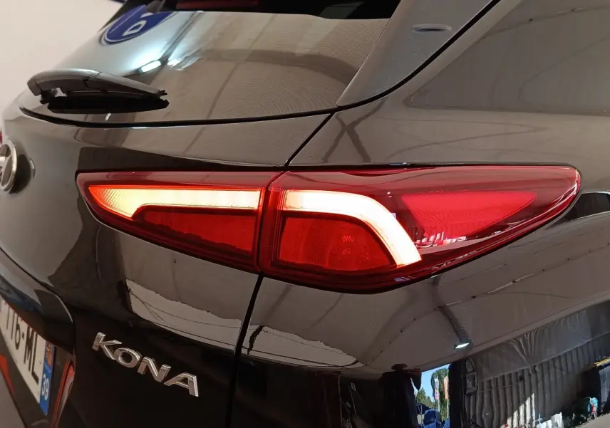 Gros plan sur le feu arrière droit allumé d'une Hyundai Kona électrique noire, avec logo et reflet d'atelier visible.