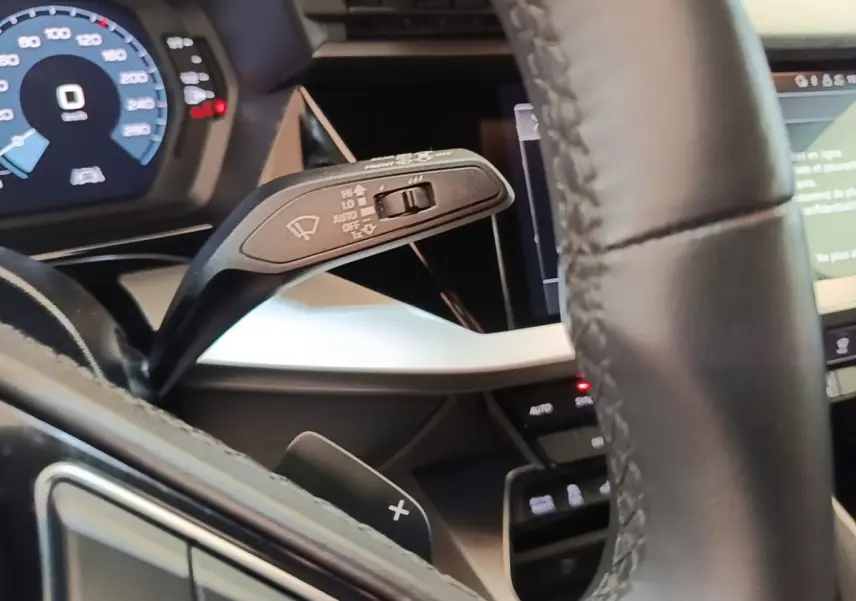 Gros plan sur le volant cuir noir et le commodo d’essuie-glace de l’Audi A3 hybride blanche, tableau de bord numérique visible.
