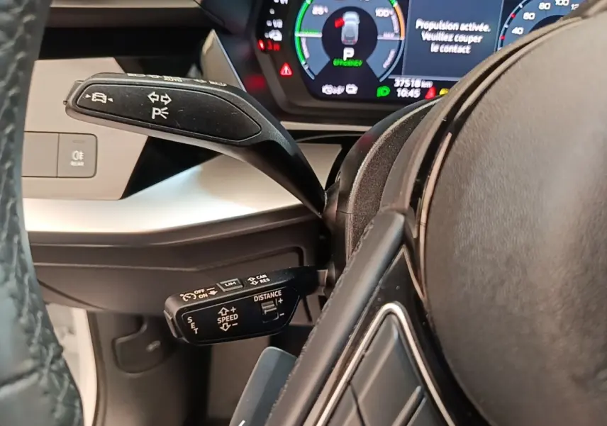 Gros plan sur le volant noir et les commandes au volant d'une Audi A3 blanche hybride rechargeable, tableau de bord digital visible.
