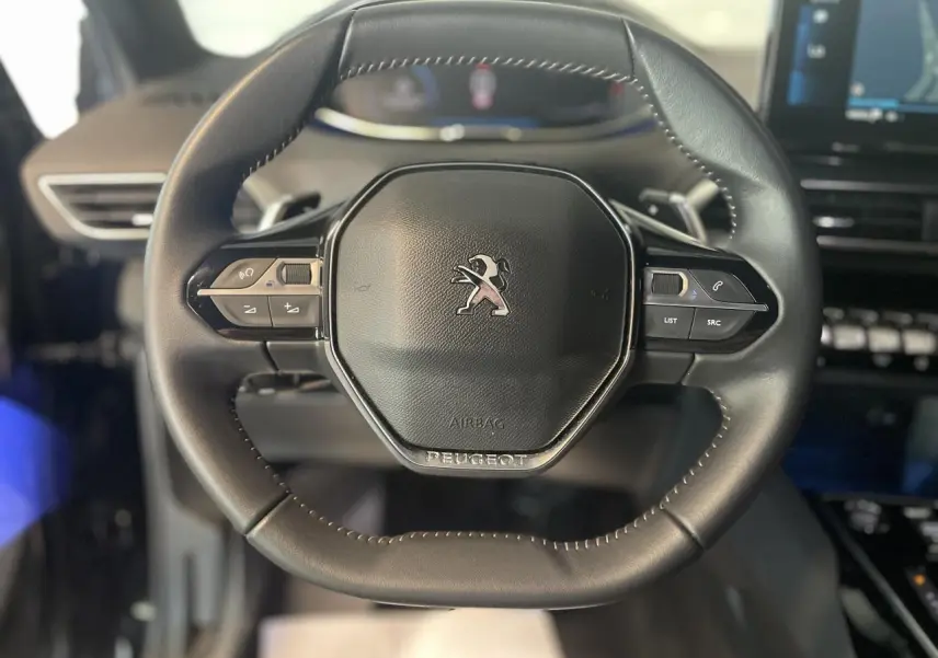 Volant compact en cuir noir avec logo Peugeot au centre, tableau de bord et écran tactile en arrière-plan flou.