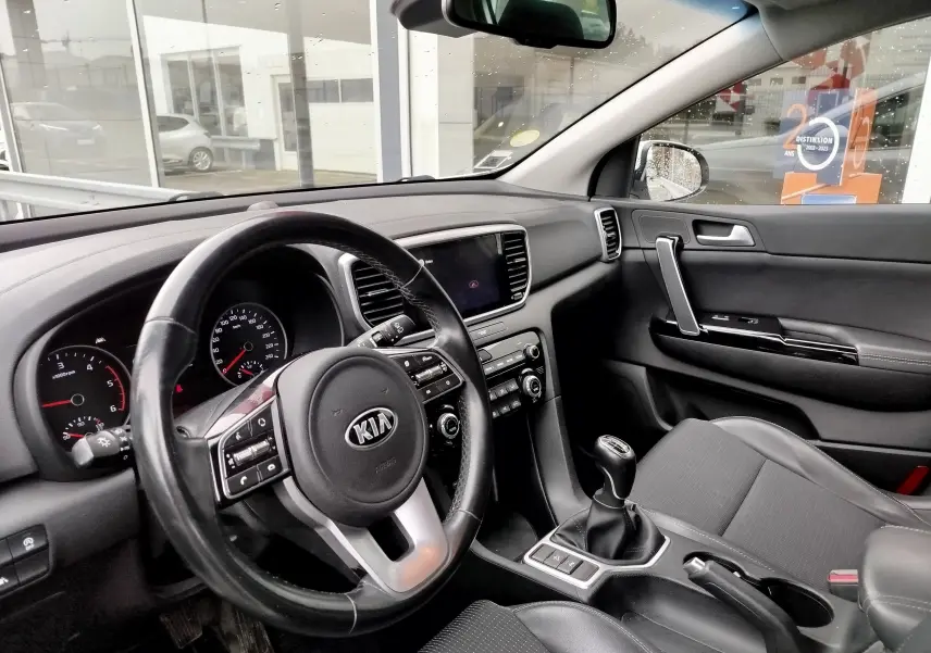 Intérieur du KIA SPORTAGE 2022 vu côté conducteur, volant cuir, tableau de bord noir et levier de vitesses manuel.
