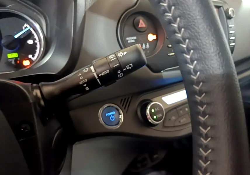 Gros plan intérieur sur le volant cuir et la commande essuie-glaces de la Toyota Yaris hybride blanche, avec bouton démarrage bleu.