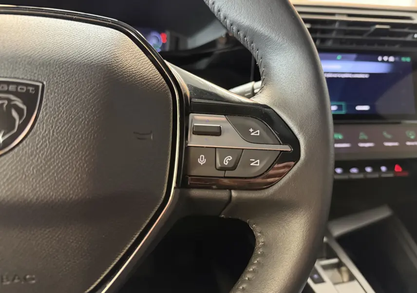 Gros plan sur le volant cuir noir de la Peugeot 308 Hybrid 145, avec commandes vocales et téléphone visibles.