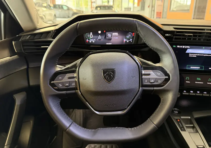 Vue rapprochée du volant cuir noir de la Peugeot 308 Hybrid 145, avec tableau de bord numérique et écran tactile visible.