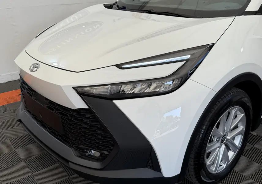 Vue 3/4 avant droit du Toyota C-HR 1.8 Hybride blanc, mettant en valeur ses phares LED et sa calandre noire texturée.