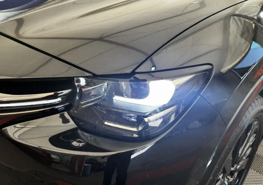 Gros plan sur le phare avant droit allumé d’un Mazda CX-60 noir, mettant en valeur la calandre et la carrosserie brillante.