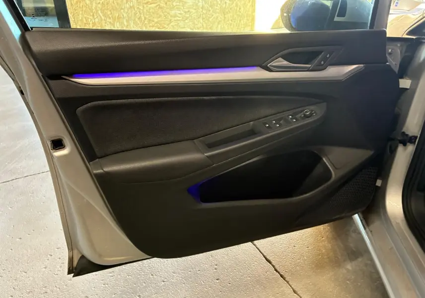 Vue intérieure de la porte avant gauche d'une Volkswagen Golf gris clair 2023, avec éclairage d'ambiance bleu et commandes électriques.