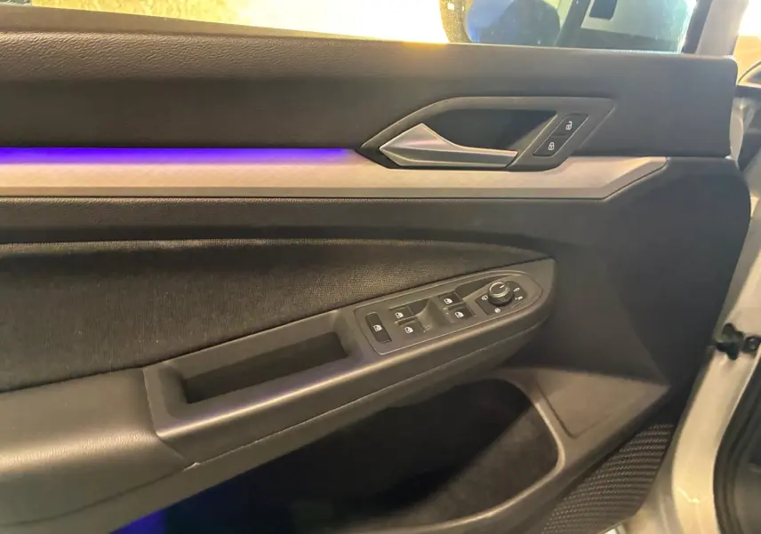 Vue intérieure côté conducteur de la porte gris clair d'une Volkswagen Golf 2023 avec éclairage d'ambiance violet et commandes électriques.