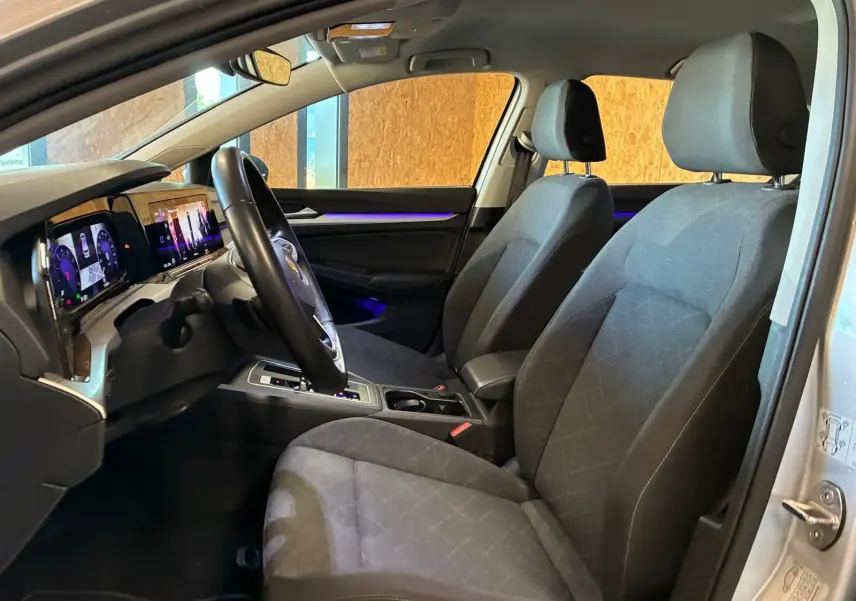 Vue intérieure côté conducteur de la Volkswagen Golf gris clair 2023, avec tableau de bord numérique et éclairage d’ambiance violet.