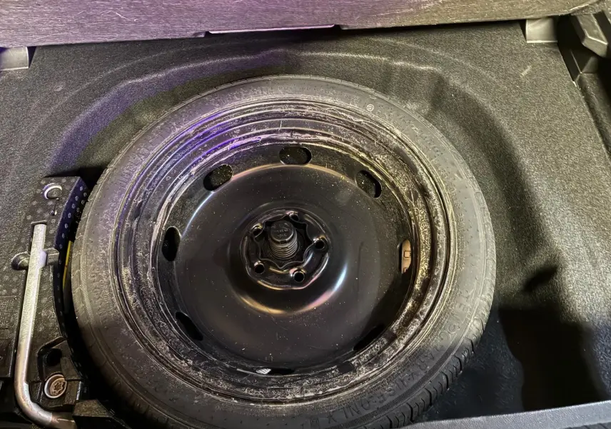 Roue de secours noire dans le coffre d’une Volkswagen Golf 2023 gris clair, vue en plongée sur le pneu et ses outils.