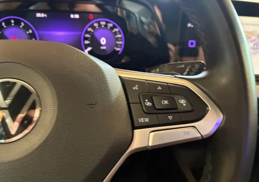 Gros plan sur le volant côté droit d’une Volkswagen Golf gris clair 2023, avec commandes intégrées et tableau de bord numérique violet.