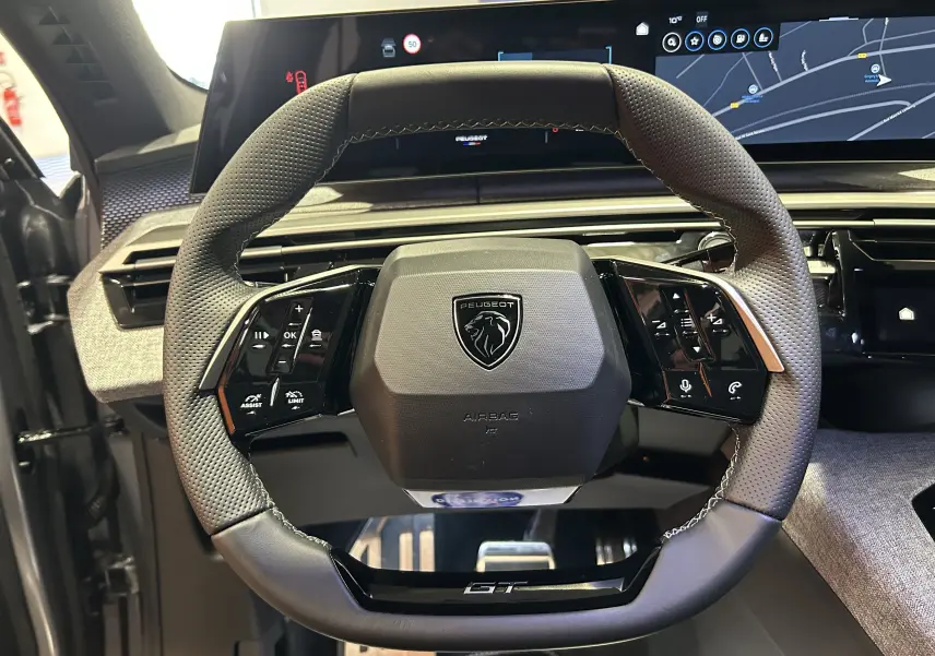 Vue rapprochée du volant GT en cuir perforé gris avec logo Peugeot, écran digital et tableau de bord du 3008 Hybrid 2025.