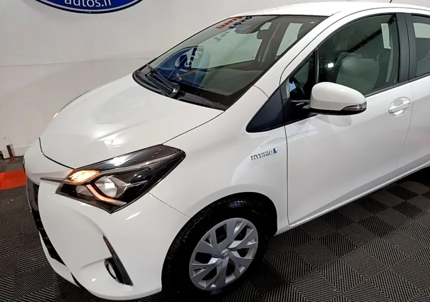 Vue 3/4 avant droit d'une Toyota Yaris blanche hybride avec phares allumés et badge Hybrid sur l'aile avant.