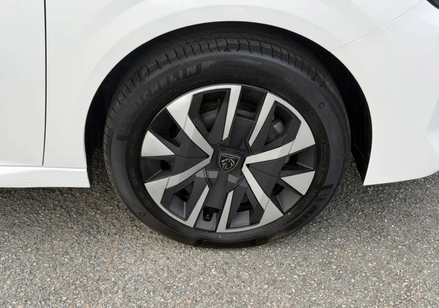 Gros plan sur la roue avant gauche blanche Okénite d’une Peugeot 208 1.2 Hybrid avec jante au design géométrique noir et argent.