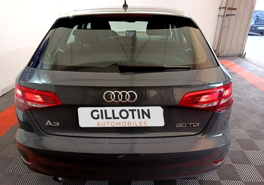 Vue arrière d'une Audi A3 30 TDI gris foncé, feux arrière allumés, dans un garage avec sol noir et rouge.