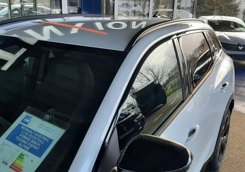 Vue 3/4 avant droit du Renault Austral blanc nacré avec toit noir et rétroviseur noir brillant, stationné devant un bâtiment.