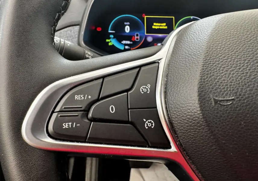 Gros plan sur les commandes du régulateur de vitesse à gauche du volant cuir noir de la Renault ZOE blanche, tableau de bord numérique en arrière-plan.