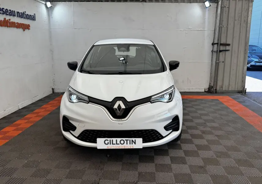 Renault ZOE Iconic R135 blanc vue de face en intérieur, phares LED allumés et calandre noire distinctive.