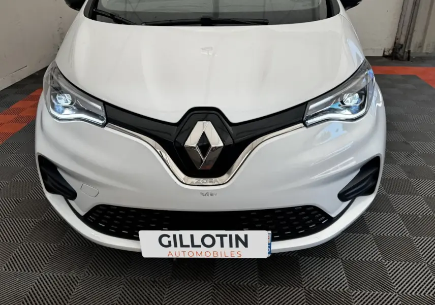 Vue avant rapprochée d'une Renault ZOE blanche avec phares LED allumés et logo Renault bien visible.