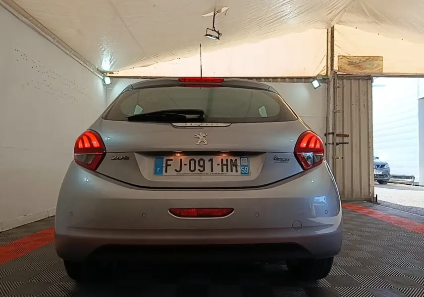 Vue arrière d'une Peugeot 208 gris clair garée en intérieur, avec feux arrière allumés et plaque d'immatriculation visible.