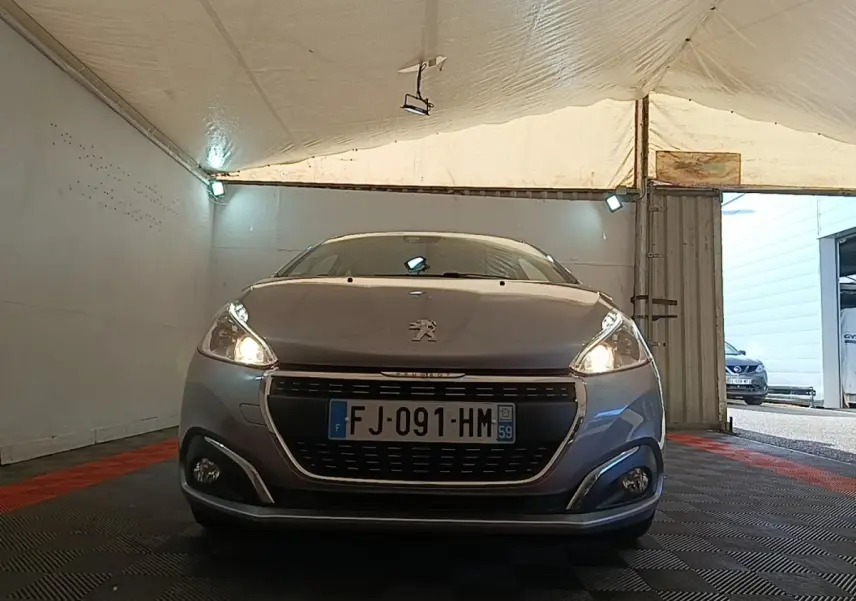 Vue frontale d'une Peugeot 208 gris clair avec phares allumés dans un garage couvert.