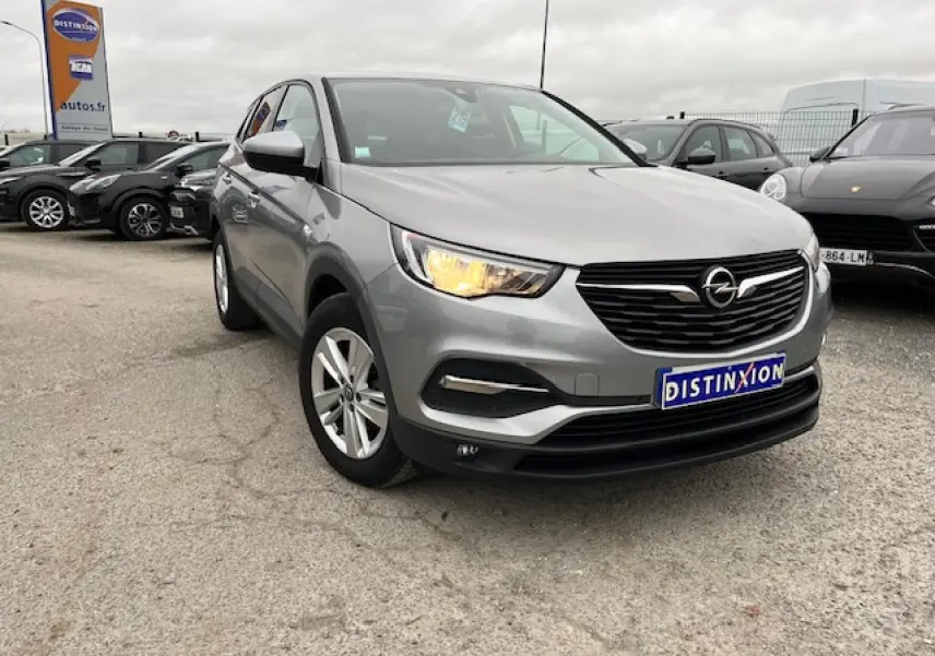 Opel Grandland X gris clair vu en 3/4 avant droit avec phare allumé et jantes alliage 17 pouces sur parking.