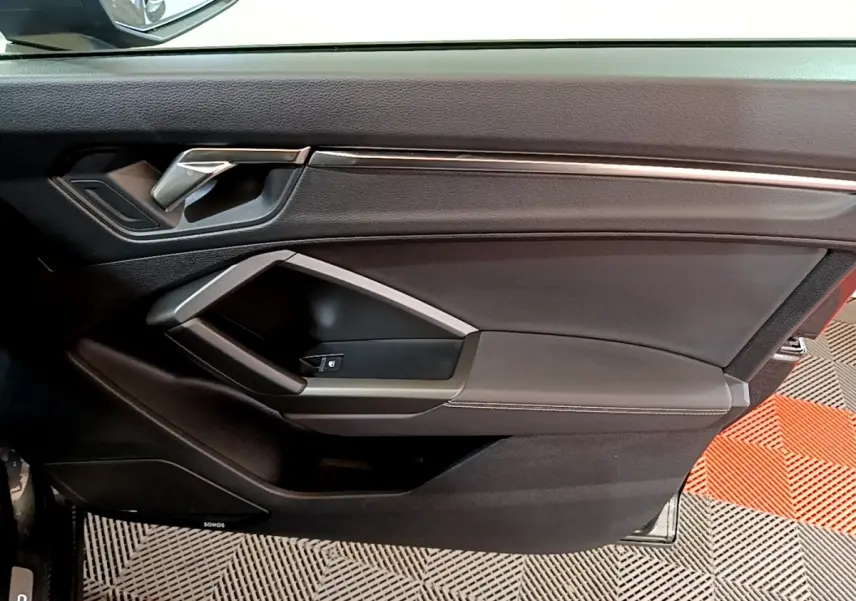Vue intérieure côté gauche de la porte avant d’un Audi Q3 gris foncé avec poignée argentée et commande de vitre.