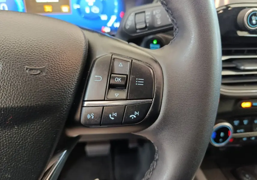 Gros plan sur les commandes gauche du volant cuir noir du Ford Kuga 2023, avec tableau de bord numérique bleu en arrière-plan.