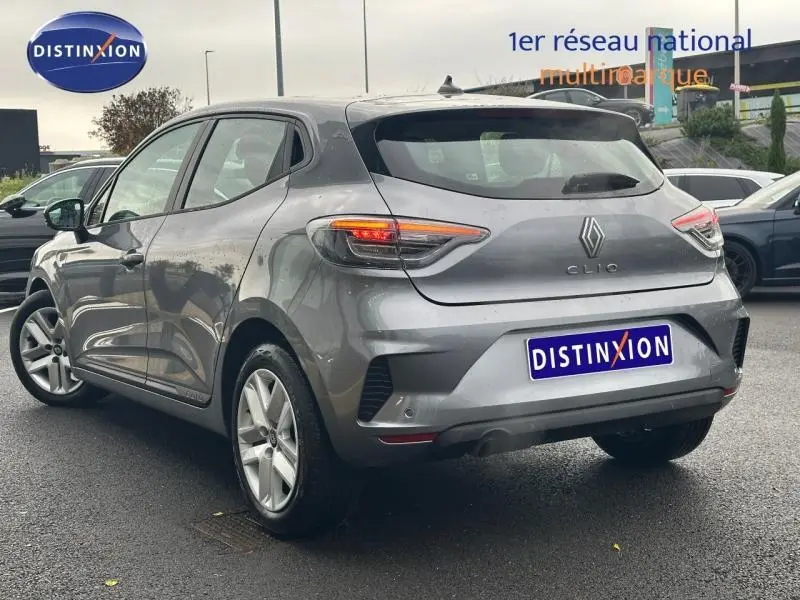 Vue 3/4 arrière droite d'une Renault Clio 1.0 TCe 90ch Evolution 2025 grise avec feux arrière allumés sur parking.