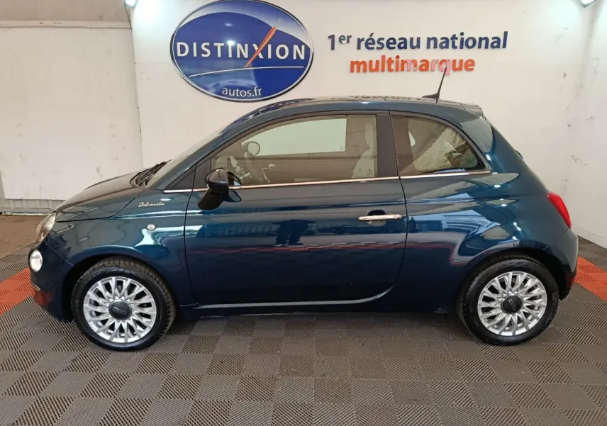 Profil côté gauche d'une Fiat 500 Dolcevita hybride bleu foncé avec jantes alliage et toit noir en intérieur showroom.
