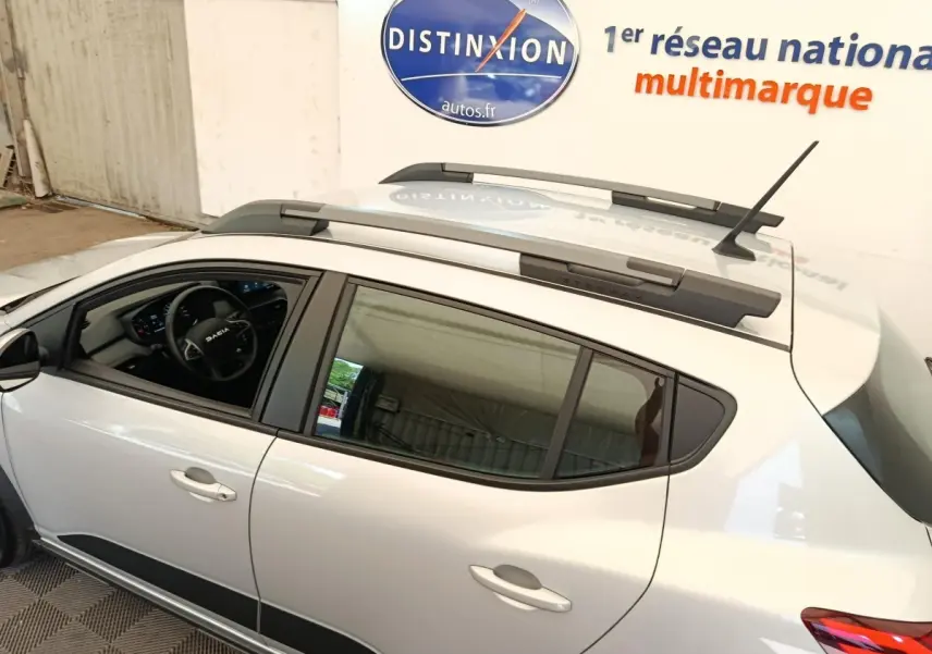 Vue latérale gauche d'une Dacia Sandero gris clair 2023 avec barres de toit et intérieur visible au volant multifonctions.