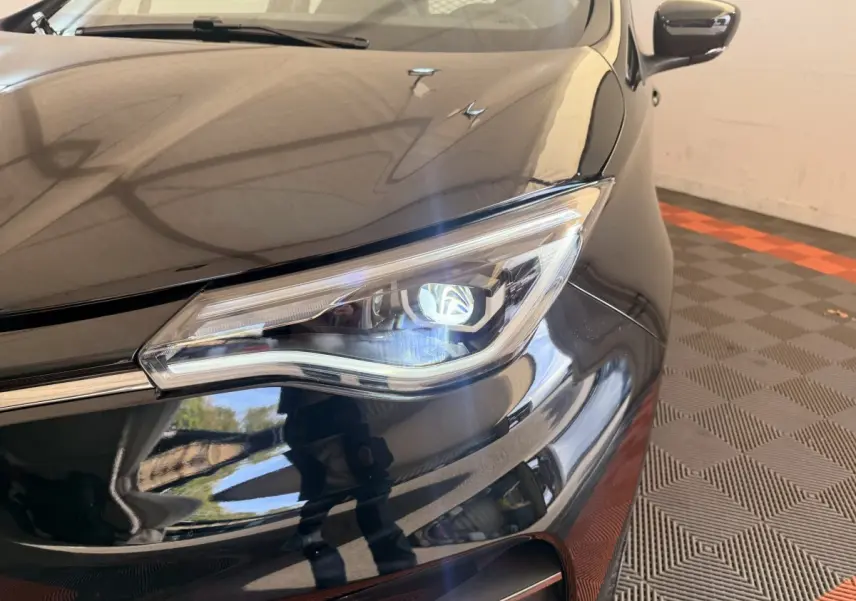 Gros plan sur le phare avant droit d'une Renault ZOE noire 2024, avec reflet brillant sur la carrosserie.