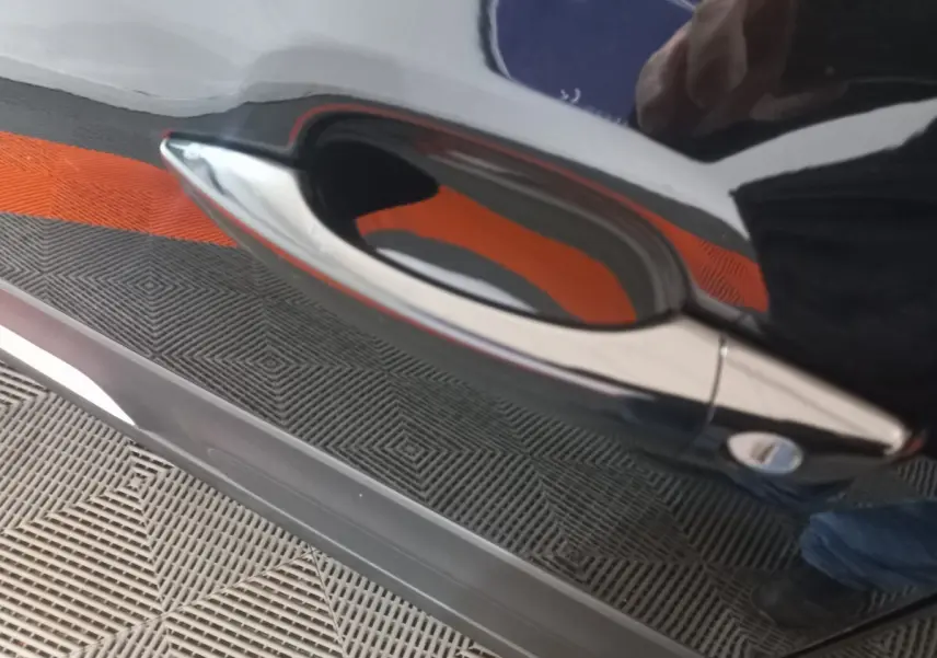 Gros plan sur la poignée de porte chromée d'une Citroën C3 noire, reflet orange et sol texturé visible.