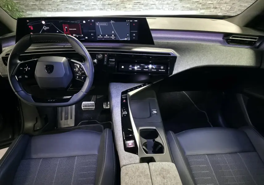 Vue intérieure avant du Peugeot 5008 2025, tableau de bord avec écran panoramique et volant compact noir, ambiance tissu gris et éclairage violet.