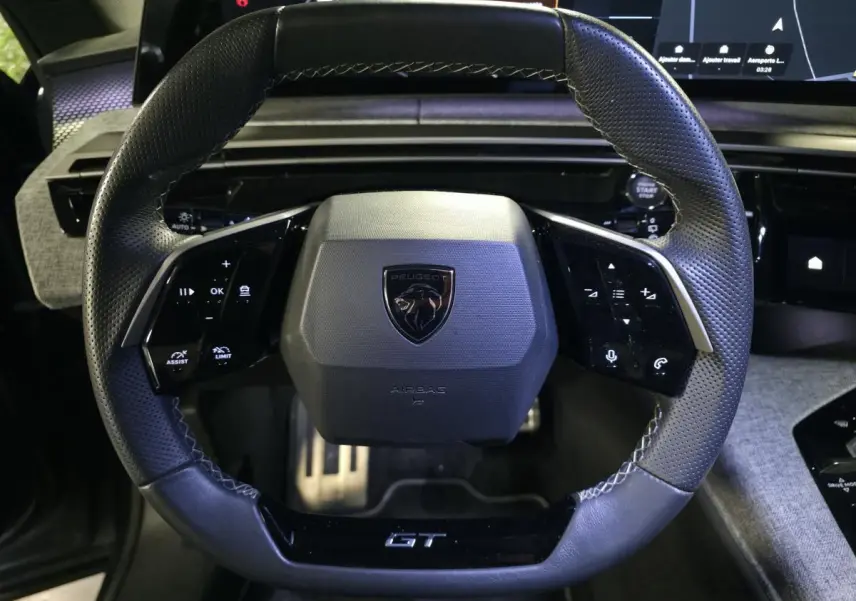 Vue rapprochée du volant noir perforé Peugeot 5008 GT avec commandes intégrées et logo central sur fond de tableau de bord numérique.